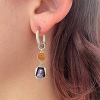 L'autre boucle d'oreille portée en gros plan sur l'oreille de la modèle. C'est la même créole en argent que l'autre boucle, mais cette fois-ci deux petits médaillons violets et oranges sont suspendus. On voit le détails de motifs religieux dans chacun d'eux.