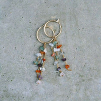 La paire de boucles d'oreilles est posée sur un fond texturé urbain en béton. On voit les deux créoles plaquées or avec chacune ses trois chaînes avec les pierres suspendues.