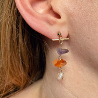 La modèle porte sur son oreille la deuxième boucle d'oreille upcyclée. Elle est aussi pendante, avec de bas en haut, une cornaline pâle, une cornaline très orangée, une améthyste, et un fermoir en T plaqué or qui complète celui de l'autre boucle.