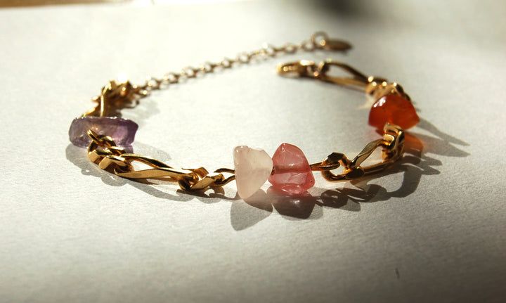 Bracelet en plaqué or constitué de pierres semi-précieuses violette, rose et orange, posé sur un fond blanc. 