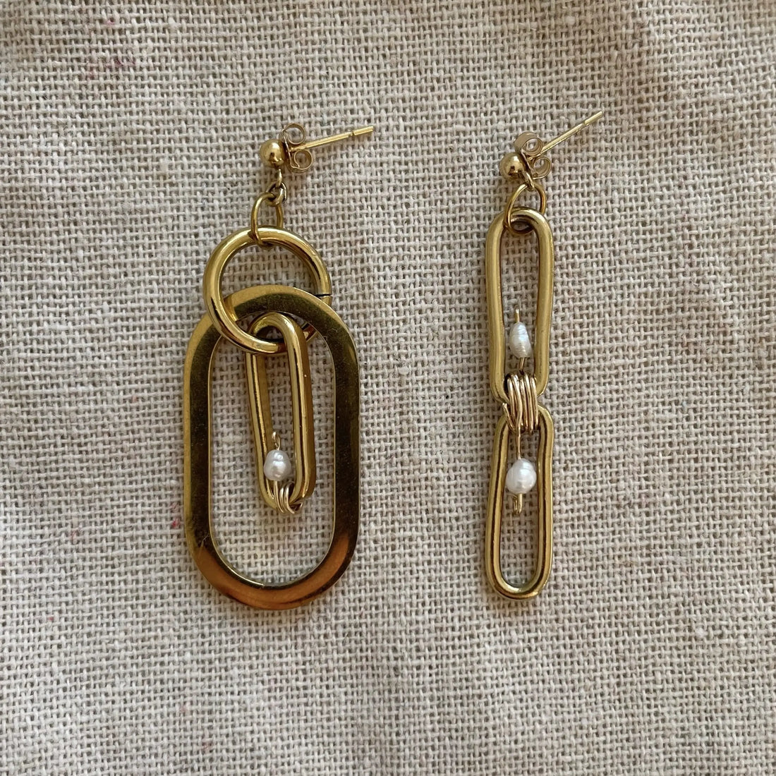 La paire de boucles d'oreilles upcyclée est posée sur un tissu beige. Elle est entièrement plaquée or avec des touches de perles d'eau douce.