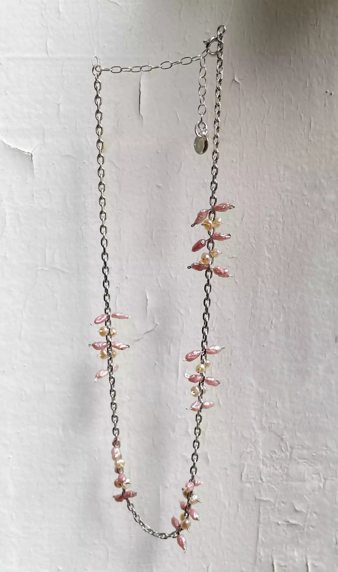 Photo du collier suspendu devant un fond blanc texturé. On voit l'alternance des motifs formés par l'assemblage des perles d'eau douce sur la chaîne en argent.