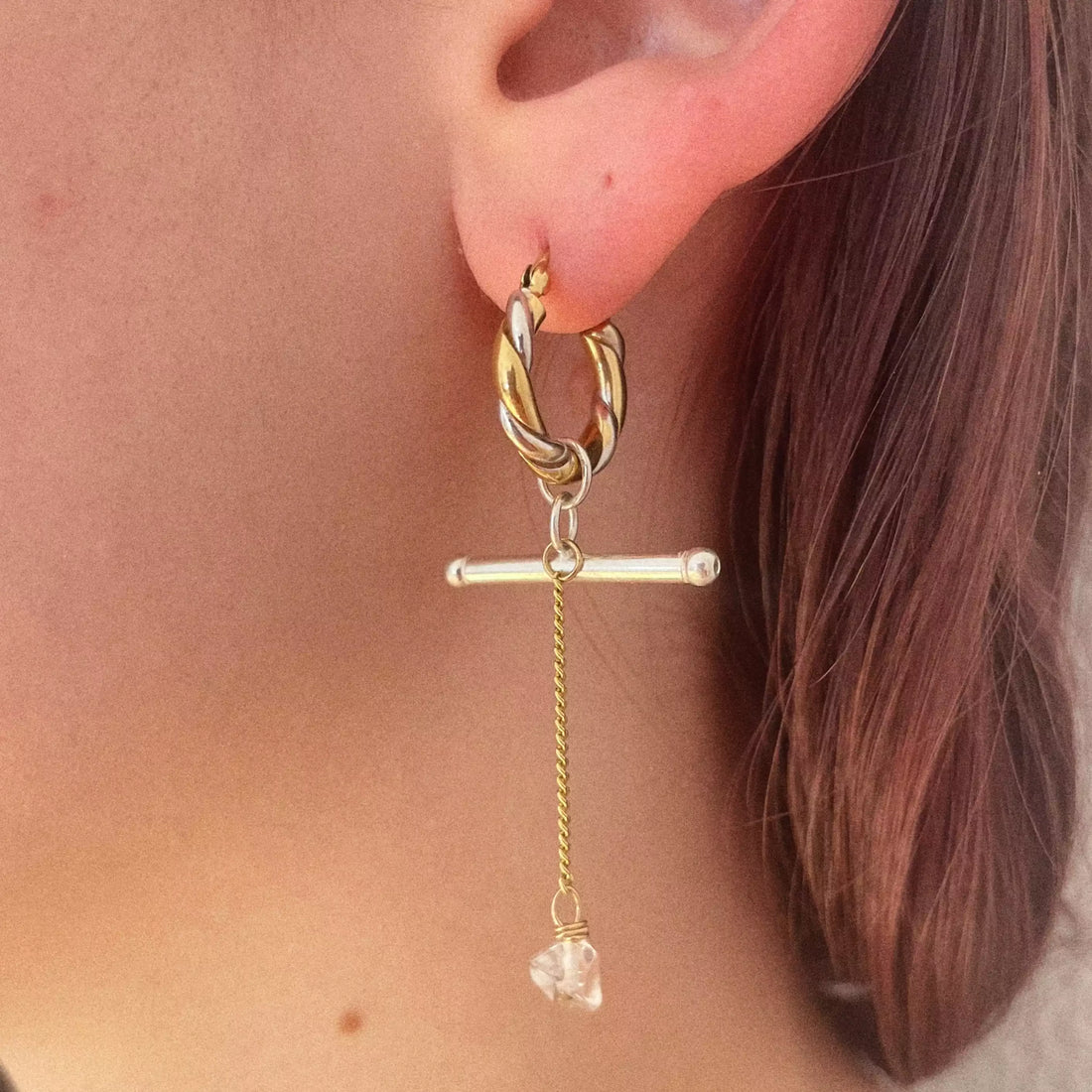 La deuxième boucle d'oreille portée. Elle est aussi pendante, avec une créoles dorée et argentée, un élément décoratif en argent, une chaîne suspendue plaquée or et un quartz transparent à l'extrémité.