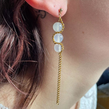 Photo portée de la première boucle, composée de trois perles de quartz blanc, entrelacées chacune par une chaîne upcyclée plaquée or, qui passe par chacune des perles en l'entourant avant de retomber pour créer un effet pendant.