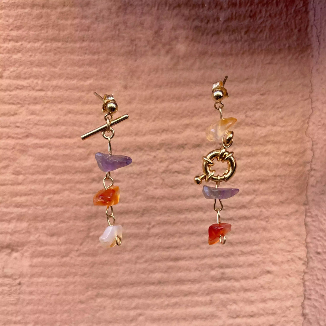 Photo de la paire de boucles d'oreilles asymétrique Tête d'Orange, sur un fond texturé en grès des vosges rose.